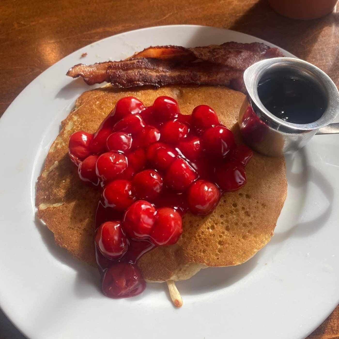 A plat with cherry-topped pancakes with syrup, and a side of bacon from Julie's Park Cafe